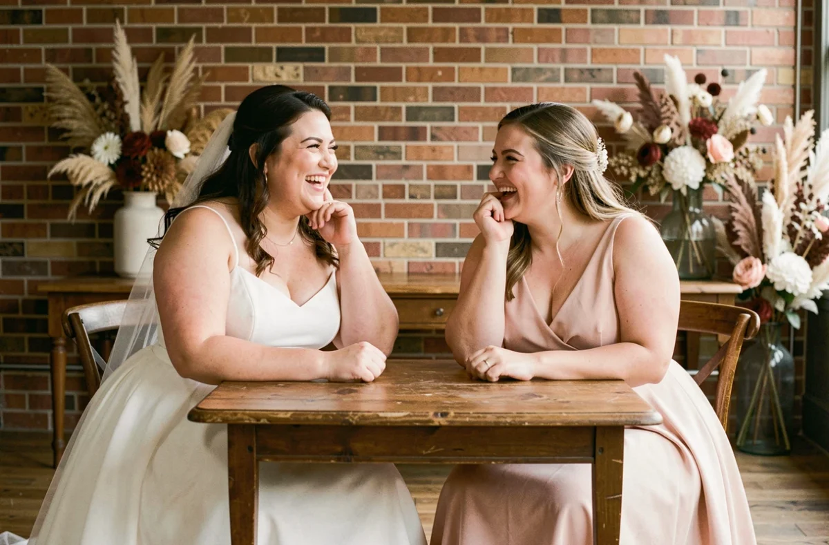 Mariée grande taille en robe empire en mousseline riant avec sa demoiselle d'honneur dans une salle décorée